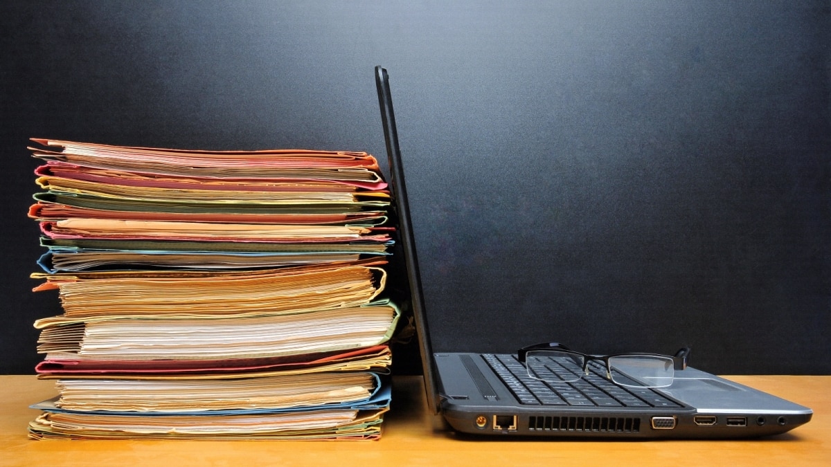 stack of papers and folders next to an open laptop with a pair of glasses on the keyboard