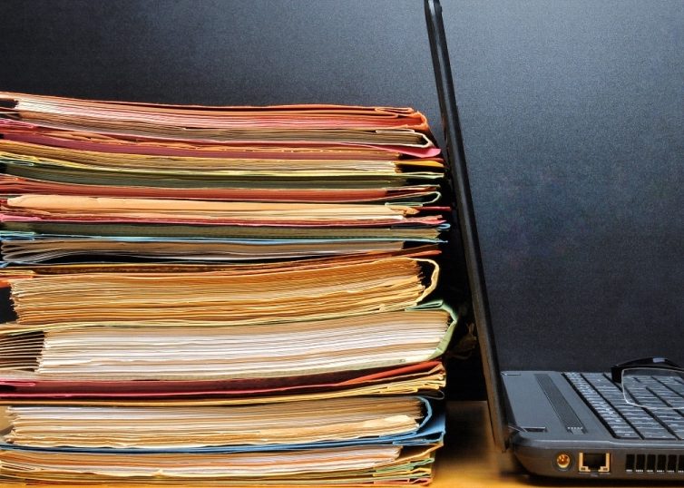 stack of papers and folders next to an open laptop with a pair of glasses on the keyboard
