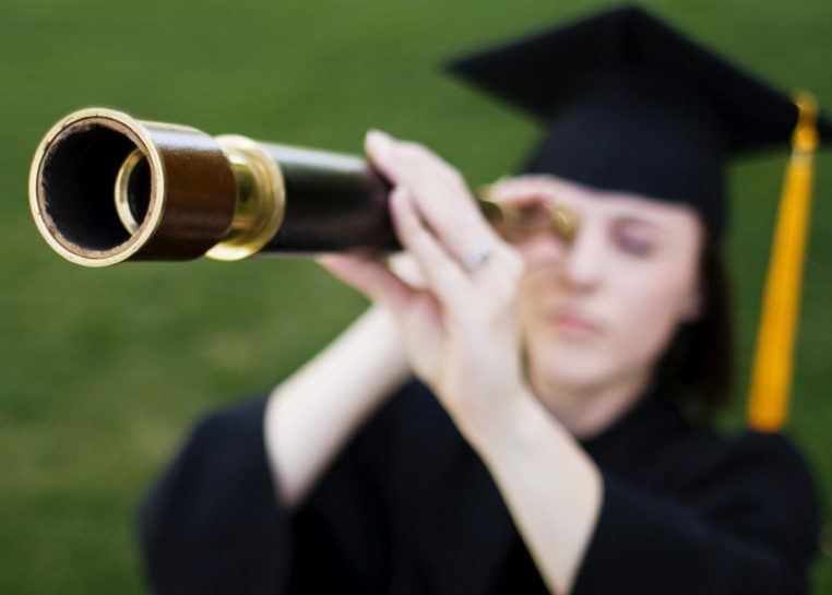Student in graduation cap and gown looking through telescope