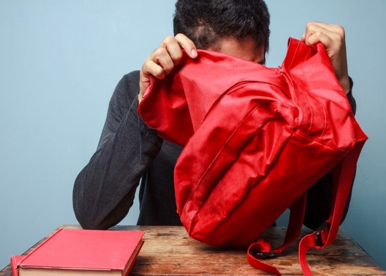 Someone sitting at a student desk with two red books on it looking into a red backpack