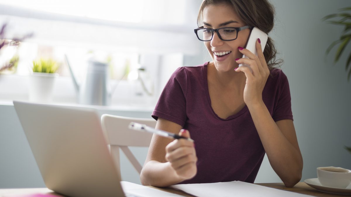 Girl holding pen and talking on a cellphone while looking at a laptop
