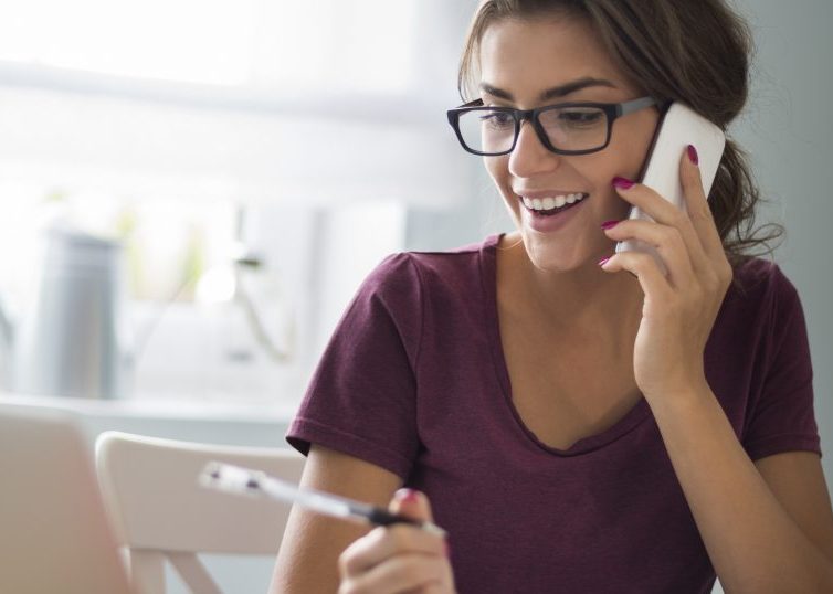 Girl holding pen and talking on a cellphone while looking at a laptop