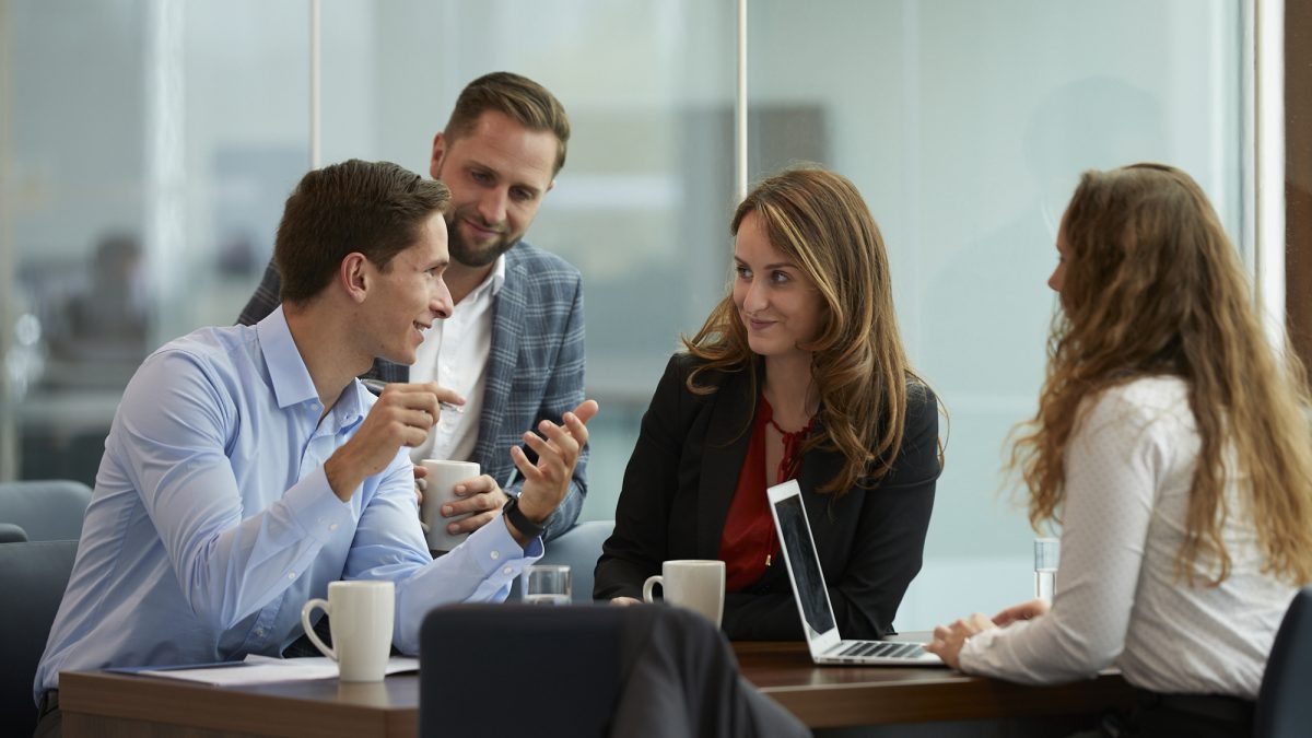 Four coworkers talking and sitting around a table with coffee mugs and a laptop