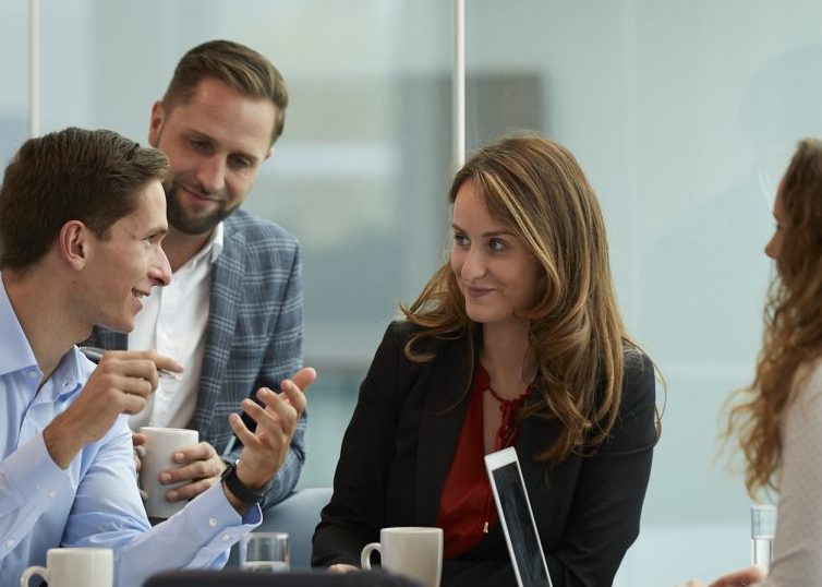 Four coworkers talking and sitting around a table with coffee mugs and a laptop