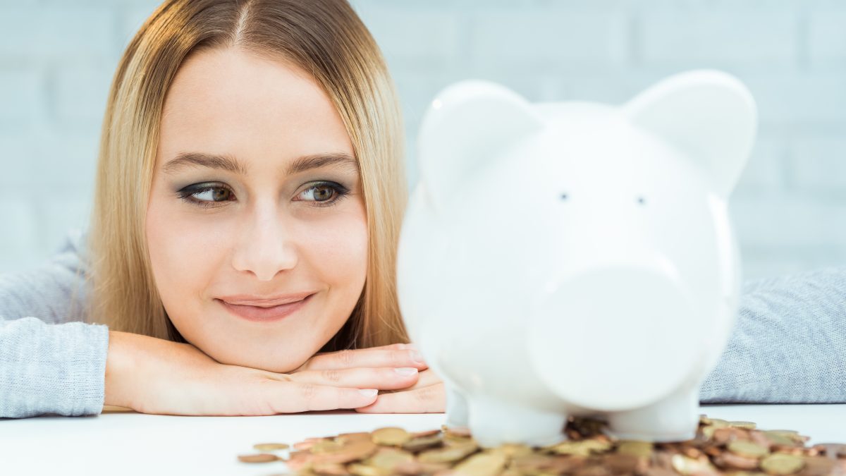 Girl smiling at white piggy bank sitting on top of pile of coins