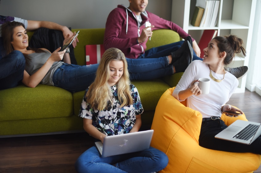 Students lounging in a living room using tablets, laptops, and cellphones