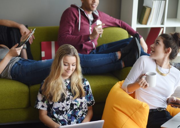 Students lounging in a living room using tablets, laptops, and cellphones