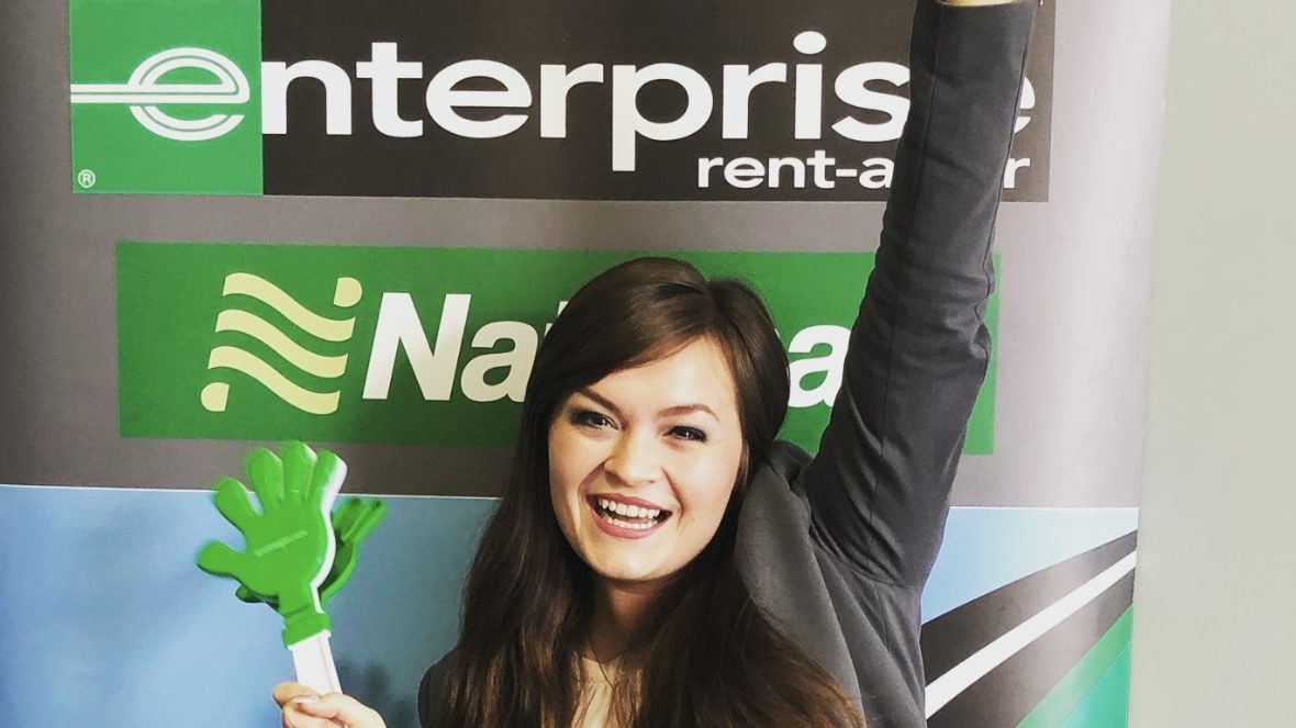 Girl holding green clapper toy in one hand with the other hand raised in front of an Enterprise sign