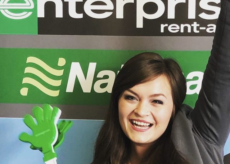 Girl holding green clapper toy in one hand with the other hand raised in front of an Enterprise sign