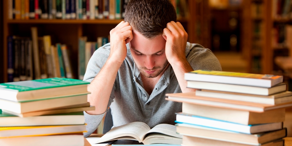 Man resting his head on his fists at a desk piled with books