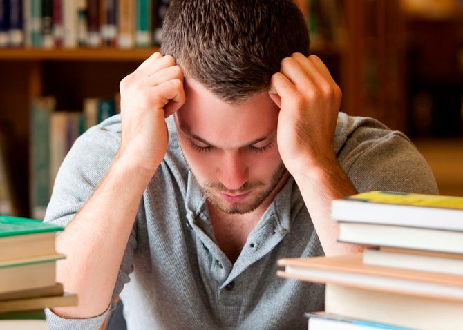Man resting his head on his fists at a desk piled with books