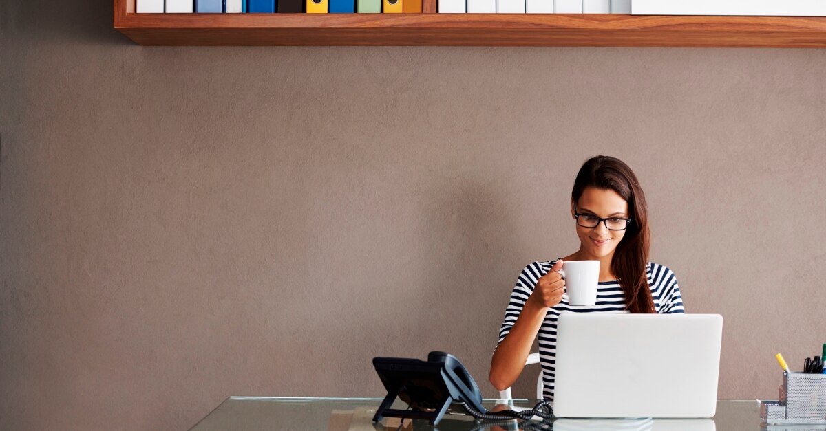 Girl sitting at desk with a laptop and phone while drinking from mug 
