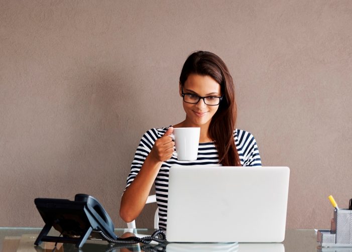 Girl sitting at desk with a laptop and phone while drinking from mug 