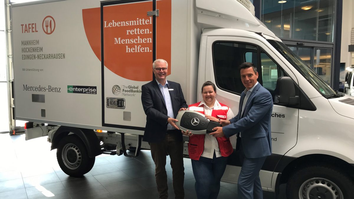 2 men in suits hand a lady in a red vest a giant Mercedes Benz key in front of a moving van