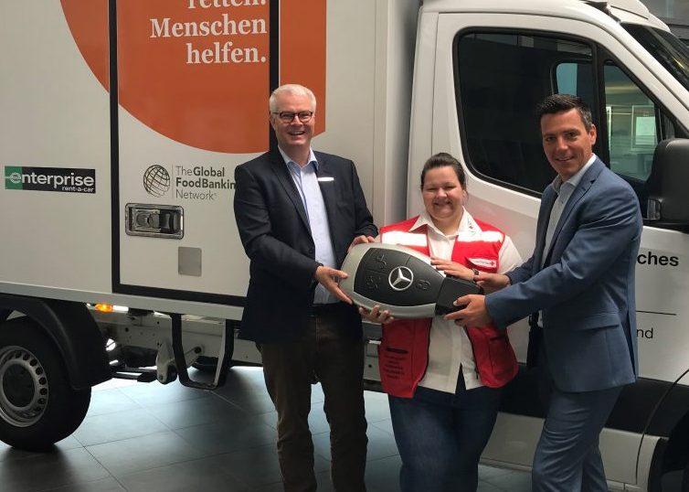 2 men in suits hand a lady in a red vest a giant Mercedes Benz key in front of a moving van