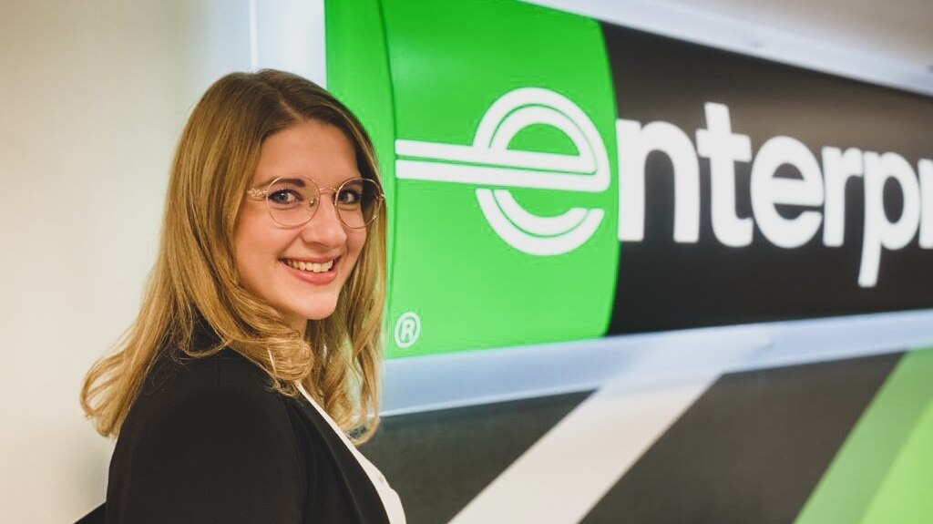 Girl with glasses smiling in front of Enterprise sign