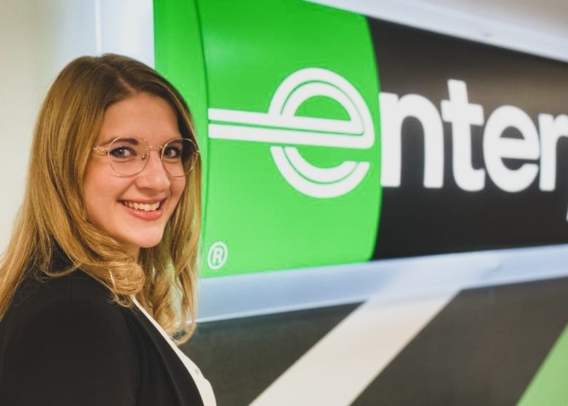 Girl with glasses smiling in front of Enterprise sign