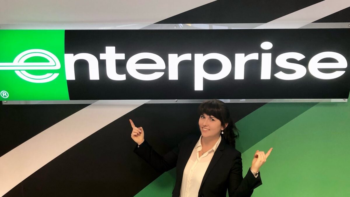 Girl in white shirt and black blazer standing underneath Enterprise sign and pointing at it with both hands