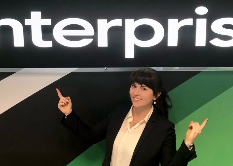 Girl in white shirt and black blazer standing underneath Enterprise sign and pointing at it with both hands