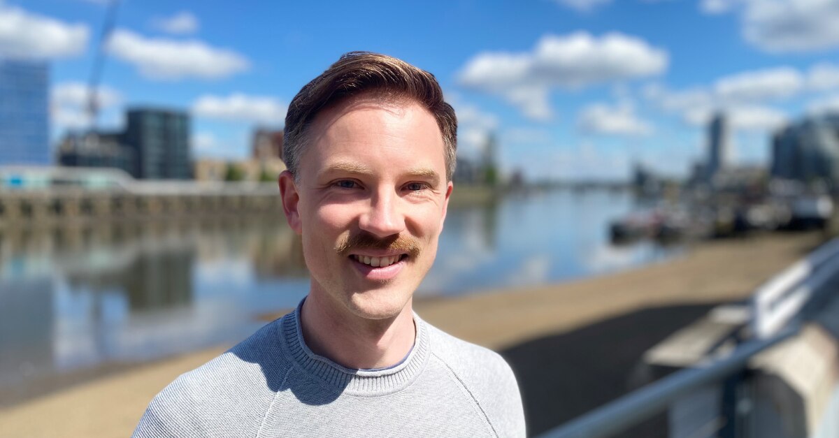 Man with mustache smiling in front of river and city