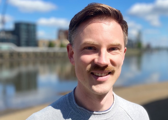 Man with mustache smiling in front of river and city