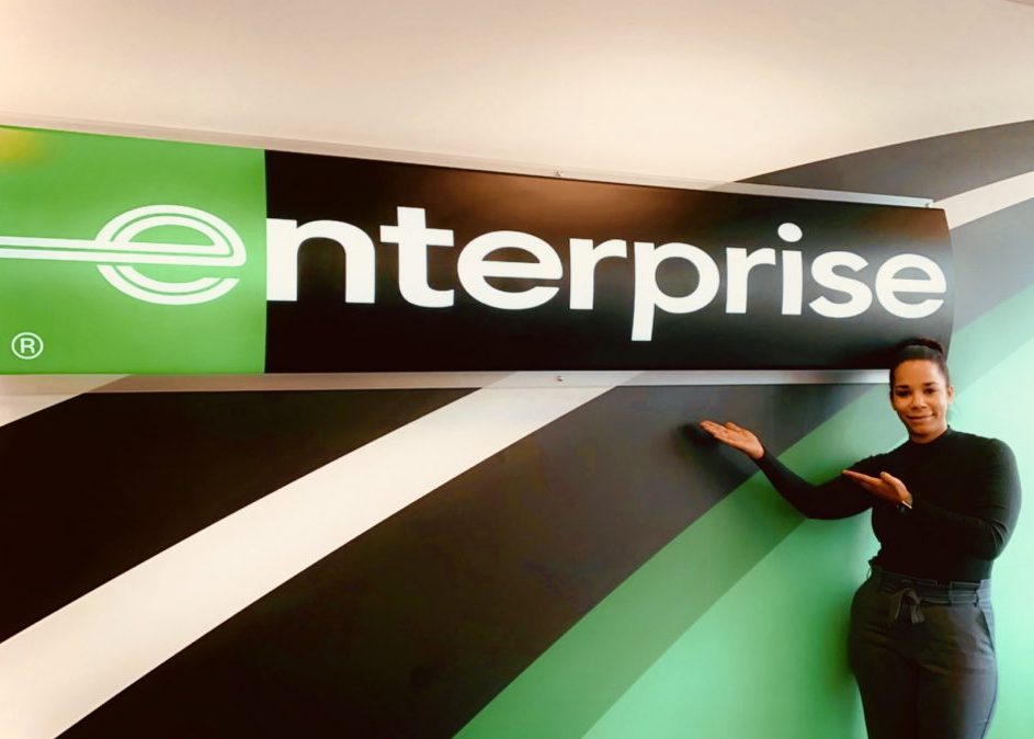 Girl wearing all black outfit showing off Enterprise sign on a wall