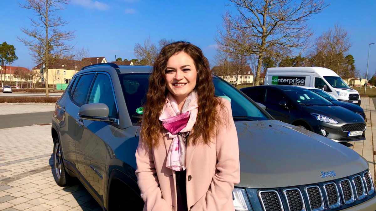 Girl in a pink coat and scarf smiling in front of a car