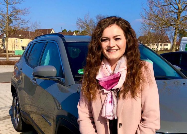 Girl in a pink coat and scarf smiling in front of a car