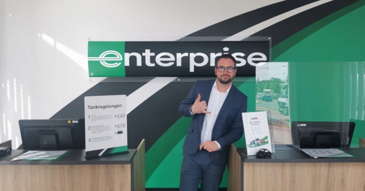 Man in suit leaning on Enterprise customer service desk giving the "hang loose" hand sign