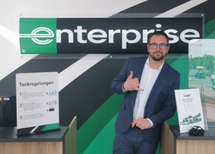 Man in suit leaning on Enterprise customer service desk giving the "hang loose" hand sign