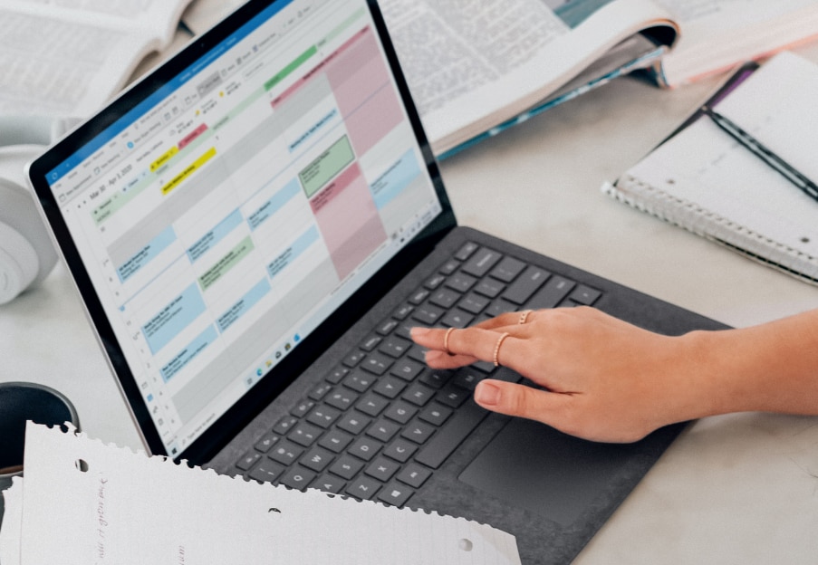 Open laptop with hand on keyboard and Outlook Calendar on screen, sitting on desk covered in paper and books