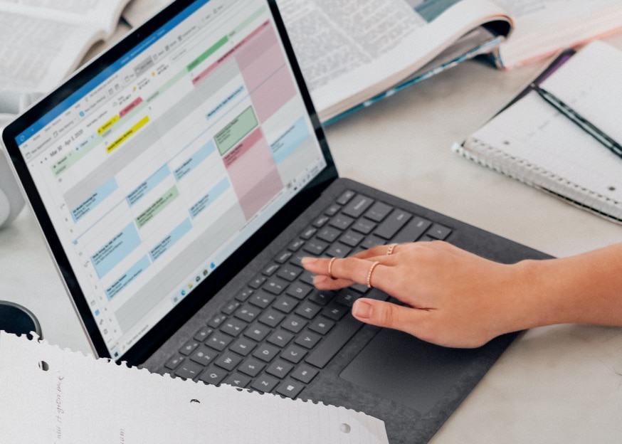 Open laptop with hand on keyboard and Outlook Calendar on screen, sitting on desk covered in paper and books