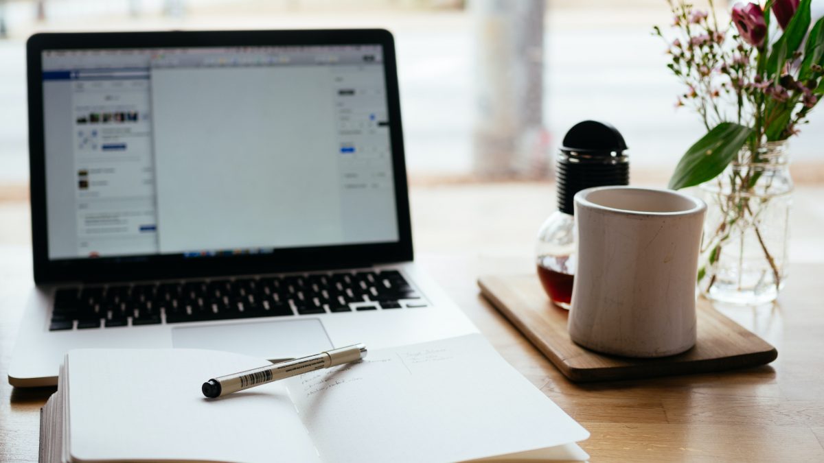 Laptop open on desk with open notebook, pen, mug, and flower vase