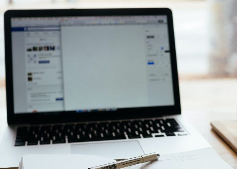 Laptop open on desk with open notebook, pen, mug, and flower vase