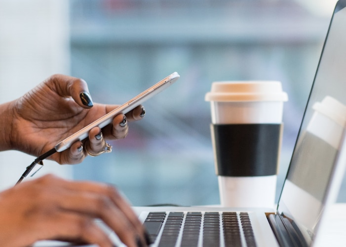Hand typing on laptop keyboard while other hand uses cellphone with a to-go coffee cup sitting next to the laptop